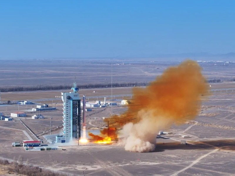 A Long March 2C rocket lifts off from Jiuquan Satellite Launch Center in China’s Gobi Desert, trailing a thick plume of orange-brown exhaust and dust as it rises beside the launch tower under a clear blue sky, with low buildings and flat desert terrain stretching into the distance.