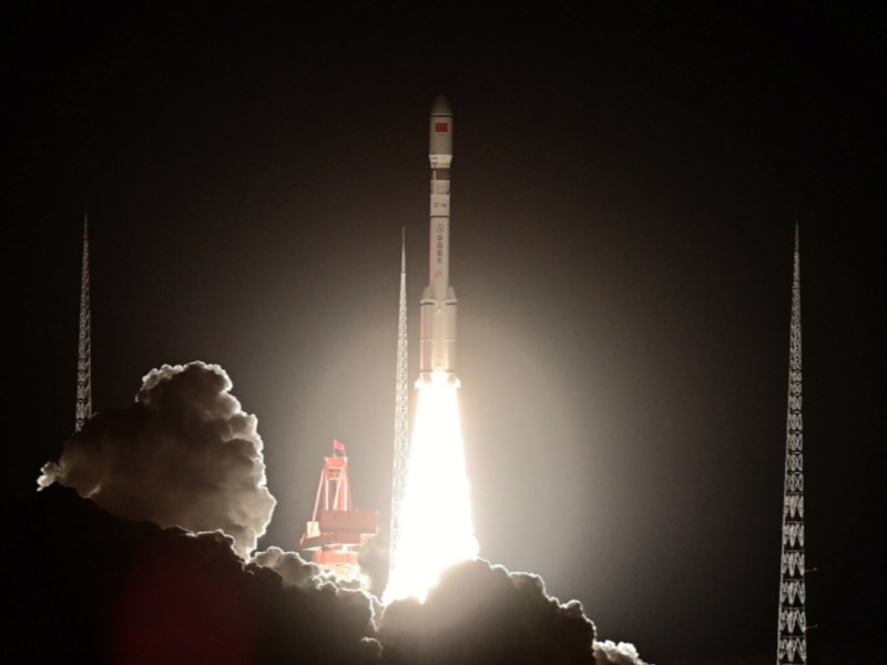 A Long March 6A rocket lifts off from a launch pad at night, its engines producing a bright plume of flame and smoke as it ascends between two tall support towers.