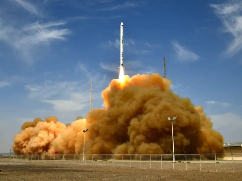 A Hyperbola-1 solid-fueled rocket developed by China's iSpace lifts off from the Jiuquan Satellite Launch Center on July 29, 2025, trailing a large plume of brownish exhaust against a clear blue sky. The rocket ascends vertically as launch gantries and fencing are visible on the desert ground below.