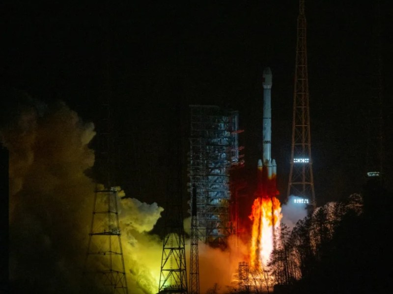 A Long March 3B rocket lifts off from Xichang Satellite Launch Center at night, carrying the ChinaSat-10R communications satellite. The rocket's engines produce a bright orange and yellow flame, illuminating the surrounding launch infrastructure and casting a glow on the smoke and support structures. Tall transmission towers and trees are visible in the foreground.