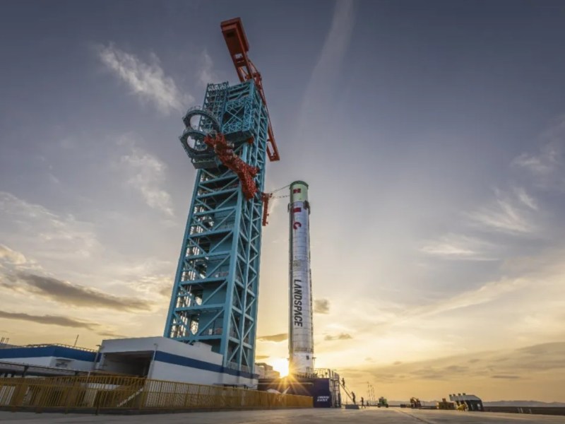 Zhuque-3 first stage stands vertically on the launch pad at Jiuquan Satellite Launch Center in northwest China, secured to a large blue service tower, with the sun setting behind it. The stainless steel rocket displays the Landspace logo prominently.