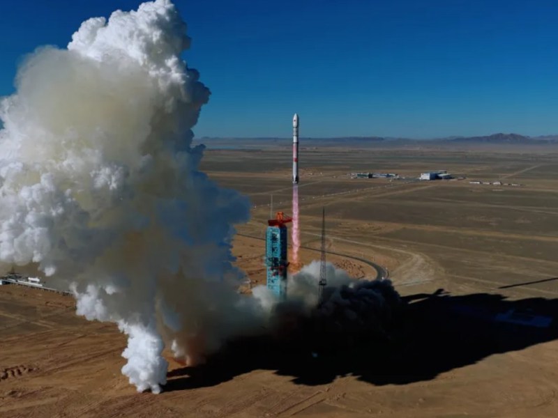 A Zhuque-3 rocket lifts off from a launch pad in a desert landscape, leaving a large plume of white exhaust behind it as it ascends into a clear blue sky. The launch tower and surrounding facilities are visible on the ground, with mountains faintly visible on the horizon.